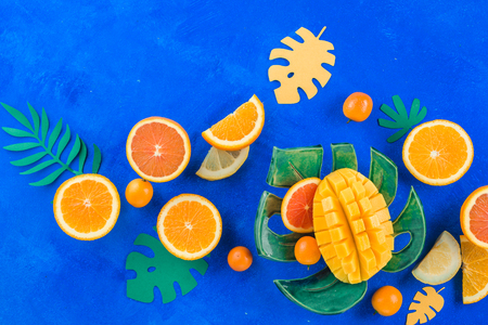Exotic fruits flat lay. Mango, oranges, kumquat and other tropical fruits on a monstera plate. Bright blue background with copy space.の写真素材