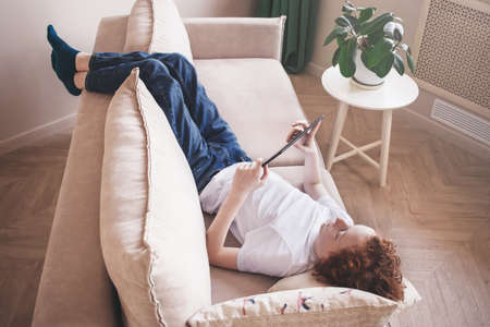 A teenager at home plays on a tablet, lies on a sofa. Toned photo.の写真素材