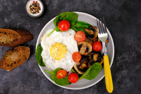 Fried egg, mushrooms, tomatoes, breads and herbs with a yellow fork on a dark background.の写真素材