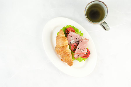 burger croissant with sausage, green, tomato, cup of green tea on white plate on a light background,...