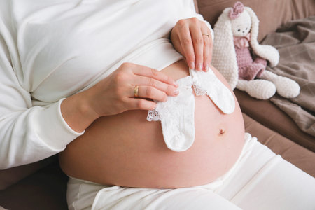Pregnant woman is holding baby socks on her belly on sofa in light living room.