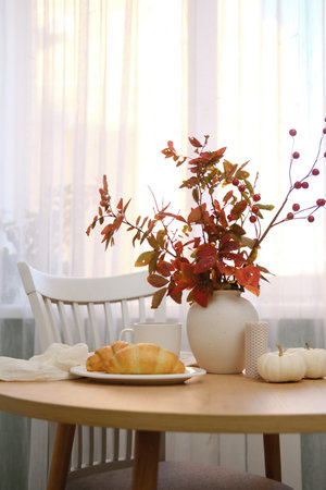 Autumn home interior. Two croissans on the plate, glass of tea, vase with autumn leaves on wooden...