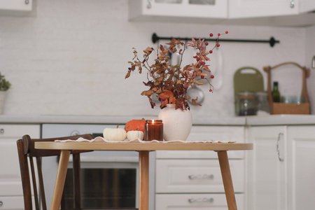 Autumn home interior- light cozy kitchen with ceramic vase and dry berries branches, burning candle, pumpkin, linen napkin on round wooden tableの写真素材