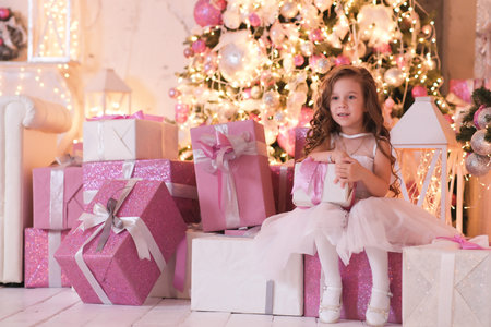 a beatiful girl with curly hair in white dress is holding of christmas box gift and sitting on gift boxes near christmas tree in light room.の写真素材
