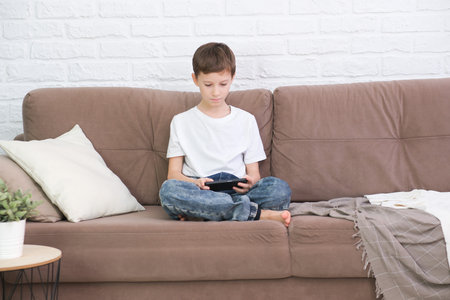 Close up cute little boy wearing headphone using smartphone, looking at screen, child holding phone in hands, sitting on couch at home alone, playing mobile device game, watching cartoons online.の写真素材