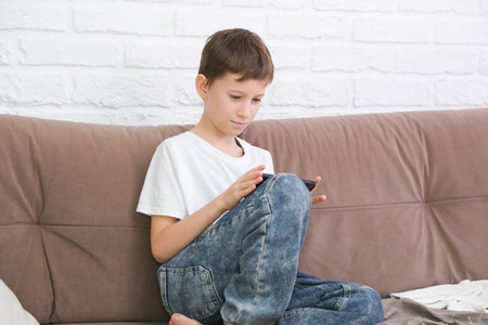 Close up cute little boy using smartphone, looking at screen, child holding phone in hands, sitting on couch at home alone, playing mobile device game, watching cartoons online.の写真素材