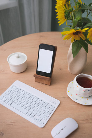 Home workplace in interior - mockup blank white screen, keyboard, computer mouse, cup of coffee, vaze with sunflowers on wooden table near the window in living room.の写真素材