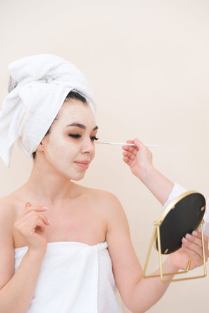 A woman is applying a facial mask while looking into a mirror and holding a towel