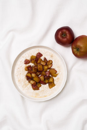 A warm bowl of oatmeal topped with caramelized apples, perfect for a cozy breakfast on white fabric...