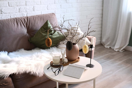 A beautifully cozy living room with modern easter home decor that includes vase with dry branches and eggs, notebook, glasses, ceramic rabbit, candles on coffee table.の写真素材