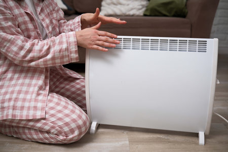 A person dressed in comfortable pajamas gently warms their hands by an electric heater at home on a chilly winter day.の写真素材