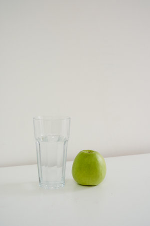 A refreshing green apple, a clear glass of water on a white table, perfect for a healthy lifestyle.の写真素材