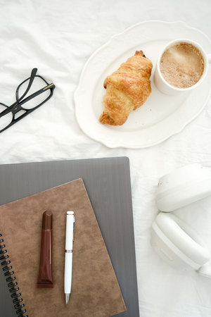 A serene scene with delicious coffee, a flaky croissant, laptop, notebook, pen, lipstick, headphones, glasses on white fabric.の写真素材