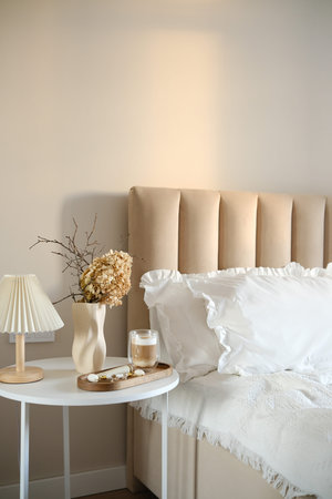 A calm and serene morning bedroom scene displays a chic bedside white round table complete with an elegant dry branches and flowers, a refreshing coffee drink, wooden tray.の写真素材
