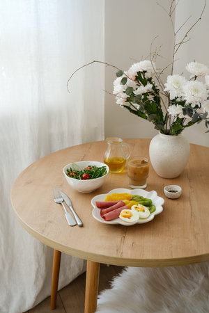 A nutritious and healthy, colorful breakfast plate features eggs, fruits, and a fresh salad with tomatoes, coffee with ice elegantly, vase with flowers arranged on a wooden tableの写真素材