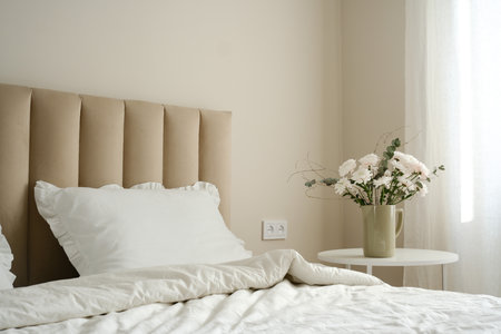A serene bedroom with a stylish bed, plush pillows, and fresh flowers on white table artfully arranged in the room.の写真素材
