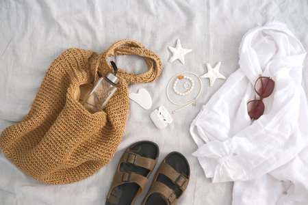 Explore stylish summer essentials for an unforgettable beach getaway, including chic attire and accessories - straw bag, linen shirt, sunglasses, shoes, earphones on white fabric.の写真素材