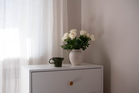 Vase with white roses with cup on dresser table in bedroom. Interior element