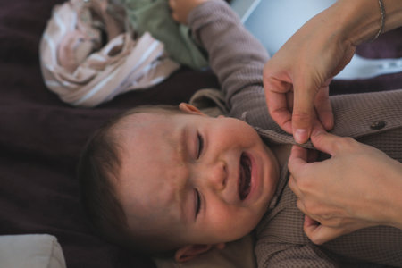 Cute baby is crying because of wearing clothes by his mother after changing him diaper. Infant boy...