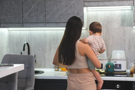 Busy Mom Holds Baby While Preparing Meal in Kitchen Tired mother struggles to cook with the baby in her armsの写真素材