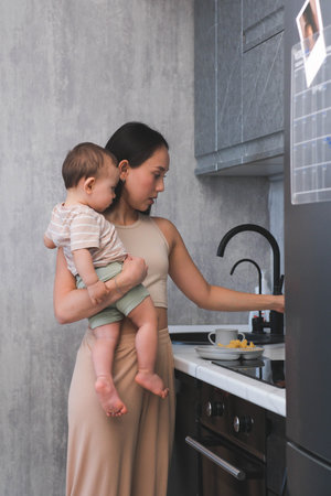 Busy Mom Holds Baby While Preparing Meal in Kitchen Tired mother struggles to cook with the baby in her armsの写真素材