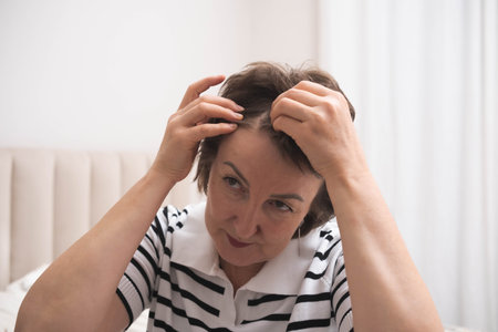 Charming woman in 50s concern look on her face checking her hair in mirror. Healthy hair, hair loss concept.の写真素材