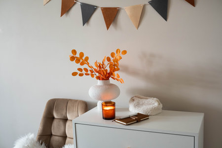 autumn decoration in bedroom. white dresser with chair. notebook, note ,candle, vase with autumn leaves and garland on wall in bedroom.の写真素材