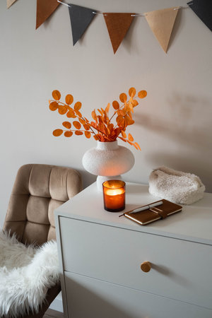 autumn decoration in bedroom. white dresser with chair. notebook, note ,candle, vase with autumn...