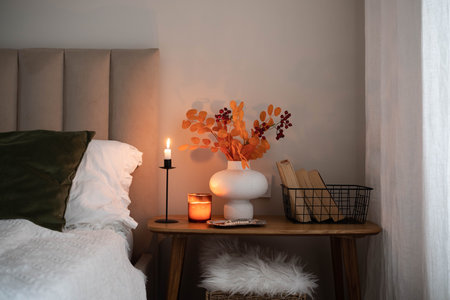 Cozy fall decor in a light and bright bedroom, bed, white bed sheets, autumn branches in white vase, candles and books on wooden bench.の写真素材