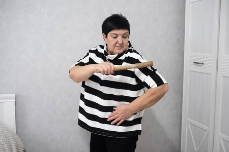 Elderly woman in a striped shirt holding a wooden massage roller across her shoulder, standing indoors and demonstrating a self-massage technique.の写真素材