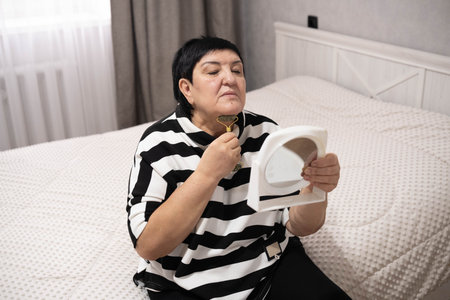 Middle-aged woman sitting on a bed in a cozy bedroom, using a facial massage roller while looking into a mirror. Self-care, skincare routine, and beauty treatment at home.の写真素材