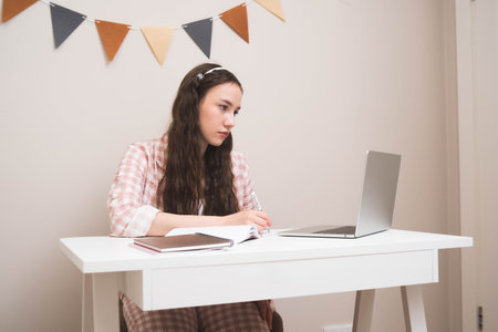 Young woman studying at a desk with a laptop and notebooks, writing notes while concentrating. Concept of online learning, education, remote work, student life, productivity.の写真素材