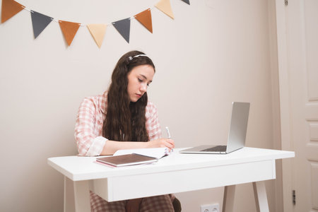 Young woman studying at a desk with a laptop and notebooks, writing notes while concentrating. Concept of online learning, education, remote work, student life, productivity.の写真素材
