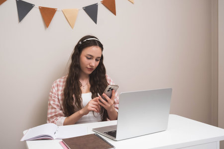 Young woman sitting at a desk with a laptop and notebooks, using her smartphone. Concept of studying, remote work, online learning, multitasking, or communication.の写真素材