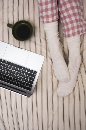 Cozy scene with a laptop, a cup of coffee, and a person in warm socks and checkered pajamas sitting on a bed. Concept of remote work, relaxation, comfort, and morning routine.の写真素材