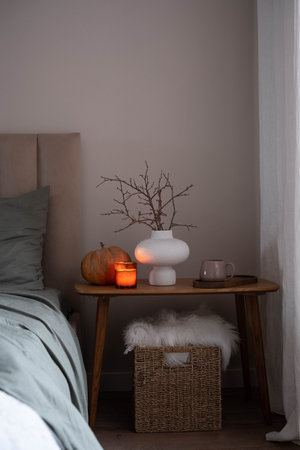 Cozy autumn bedroom corner with a pumpkin, glowing candle, ceramic vase with branches, and a mug on a wooden table, creating a warm and peaceful seasonal atmosphere.の写真素材