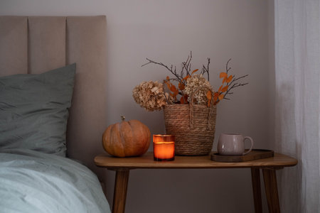 Cozy autumn bedroom corner with a pumpkin, lit candle, dried flowers in a wicker basket, and a mug on a wooden bedside table, creating a warm seasonal atmosphere.の写真素材