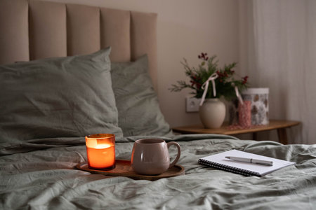 Cozy bedroom scene with a lit candle, warm mug, and notebook on a wooden tray placed on a bed with...