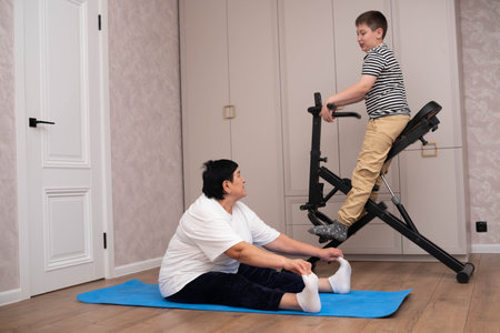 Senior woman stretching on a yoga mat at home while a child sits on an exercise machine, showing family activity, fitness, and a healthy lifestyle together.の写真素材