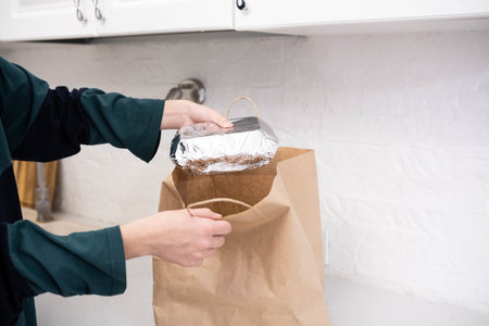Person removing a foil container from a brown paper bag on a kitchen counter. Concept of takeaway,...