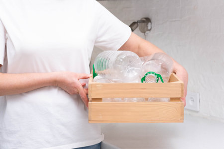 Woman in white shirt holds a wooden crate filled with plastic bottles and containers in a kitchen....