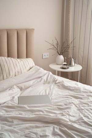 A serene bedroom scene: unmade white bed with a laptop and a small round table with a white vase of...
