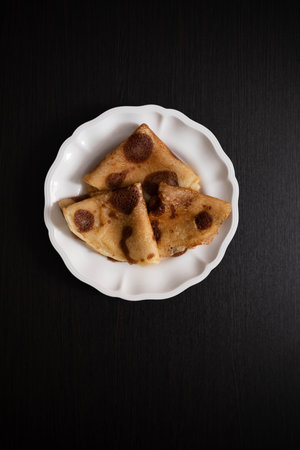 Three golden pancakes folded into triangles rest on a white scalloped plate against a dark background. The pancakes feature dark spots, showcasing their crispy texture.の写真素材