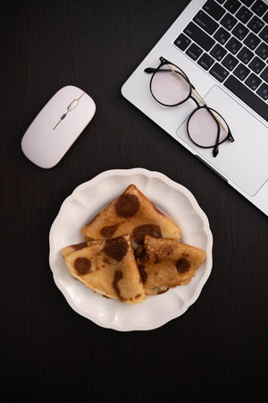 A stylish workspace scene: a plate with folded golden pancakes sits on a dark desk alongside a white...