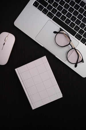 A sleek workspace setup: silver laptop with round-framed glasses on the trackpad, a white wireless mouse, and a clean grid-lined notebook on a dark desk.professional aesthetic for work or study.の写真素材