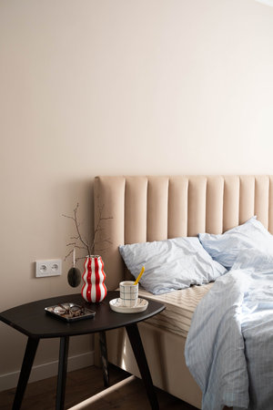 Cozy spring bedroom corner with beige tufted headboard, light blue striped bedding, black bedside...