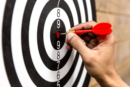 hand holding darts and stabbing or puncturing on dart board.Business target or goal success and...