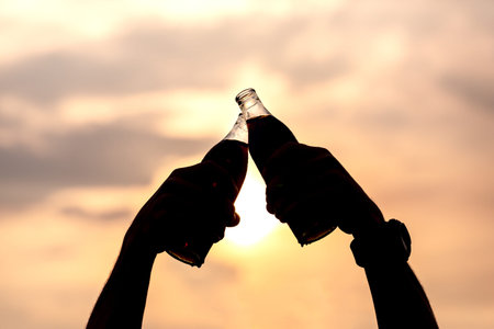 Hand of man clink soft drink bottles and raise on the sky in evening with sunlight.Silhouettes in the light of sunset.celebration and refreshment energy concept.の写真素材