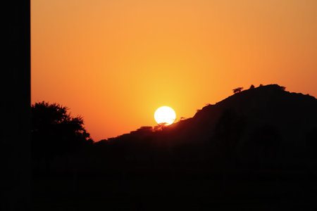 A landscape sunrise photo with a bright sun shining brightly from behind a silhouette mountain range and wild silhouette trees.concept to global warning , climate changingの写真素材