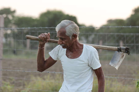 Close-up photo of An Indian senior male farmer man works in the field with a shovel on his shoulder with wearing a white vestの写真素材